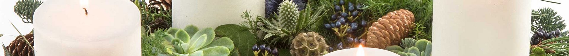 En dekorativ adventskrans med fire hvide stearinlys, der er omgivet af grønne planter, kogler og bær. Stearinlysene er tændt og skaber en hyggelig atmosfære. Kransen er fyldig og prydet med naturmaterialer.